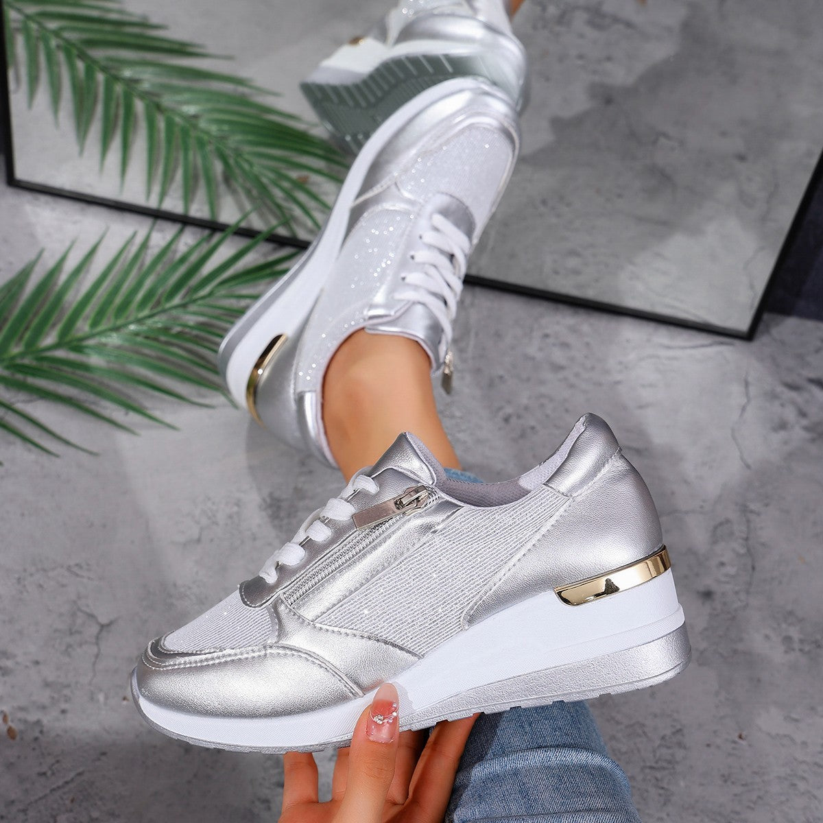 Silver sneakers with gold accents worn by a person on a gray surface with green leaves.