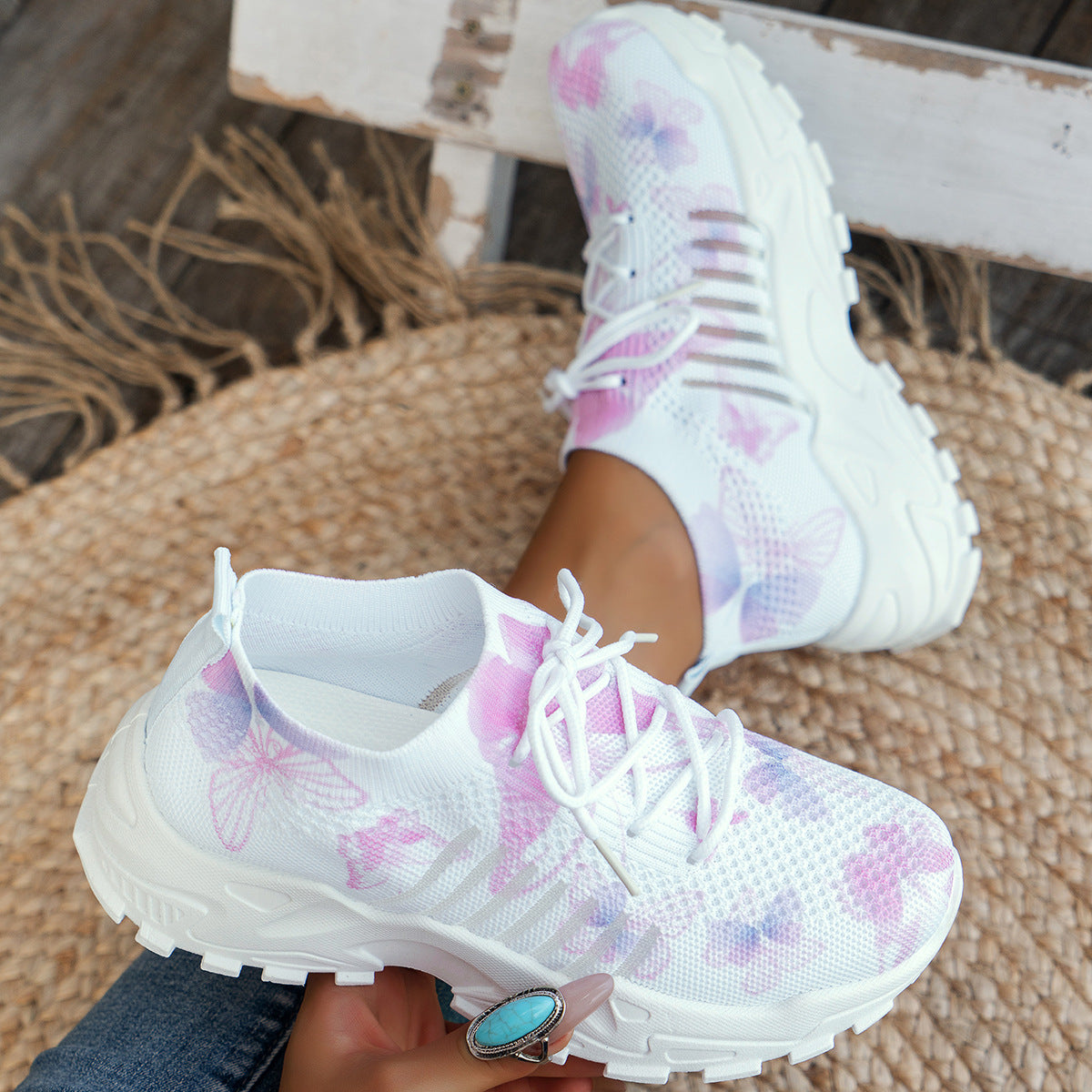 White sneakers with pink floral patterns worn by a person sitting on a woven chair.