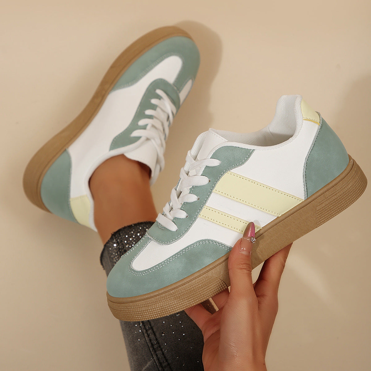 White sneakers with green and yellow accents held by a hand against a beige background